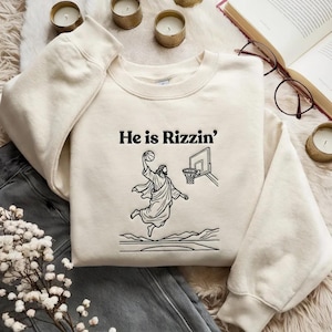 May include: Cream-colored sweatshirt with the text "He is Rizzin'" above a black and white illustration of Jesus playing basketball. The graphic depicts Jesus mid-air, dunking a basketball. The sweatshirt has long sleeves.