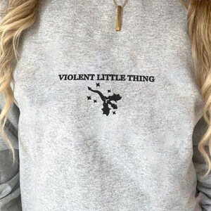 Puede incluir: Una sudadera gris jaspeada con el texto "VIOLENT LITTLE THING" y un peque&ntilde;o gr&aacute;fico negro de un drag&oacute;n. La sudadera tiene un corte holgado y cuello redondo.