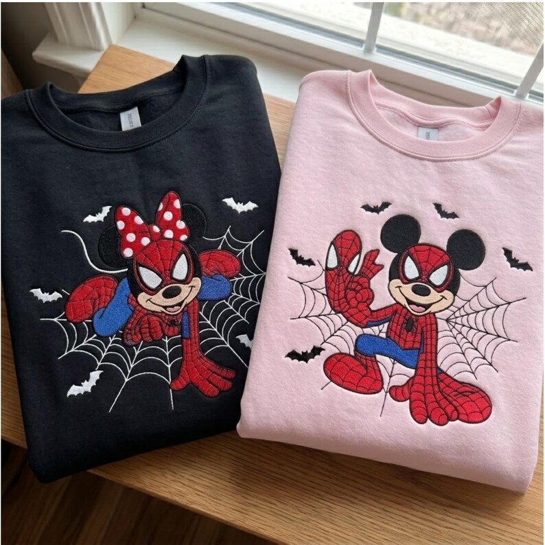 May include: A black sweatshirt and a light pink sweatshirt, each featuring an embroidered graphic of a cartoon mouse character dressed in a red and blue spider-themed costume, positioned against a spiderweb background with small black bats.