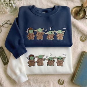 Puede incluir: Una sudadera azul marino y una sudadera blanca, cada una con un bordado de cuatro criaturas verdes con t&uacute;nicas marrones. El dise&ntilde;o incluye una criatura con auriculares, una extendiendo la mano, una con un signo de interrogaci&oacute;n y el texto coo!