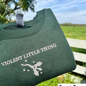 Puede incluir: Sudadera verde bosque con el texto "VIOLENT LITTLE THING" y un dise&ntilde;o de drag&oacute;n bordado en blanco.