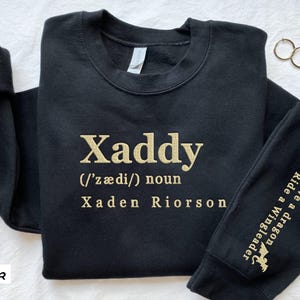 May include: Black sweatshirt with gold lettering that reads "Xaddy (/ˈz&aelig;di/) noun Xaden Riorson". The right sleeve has a gold dragon graphic and the text "Ride a Wingleader Save a dragon".