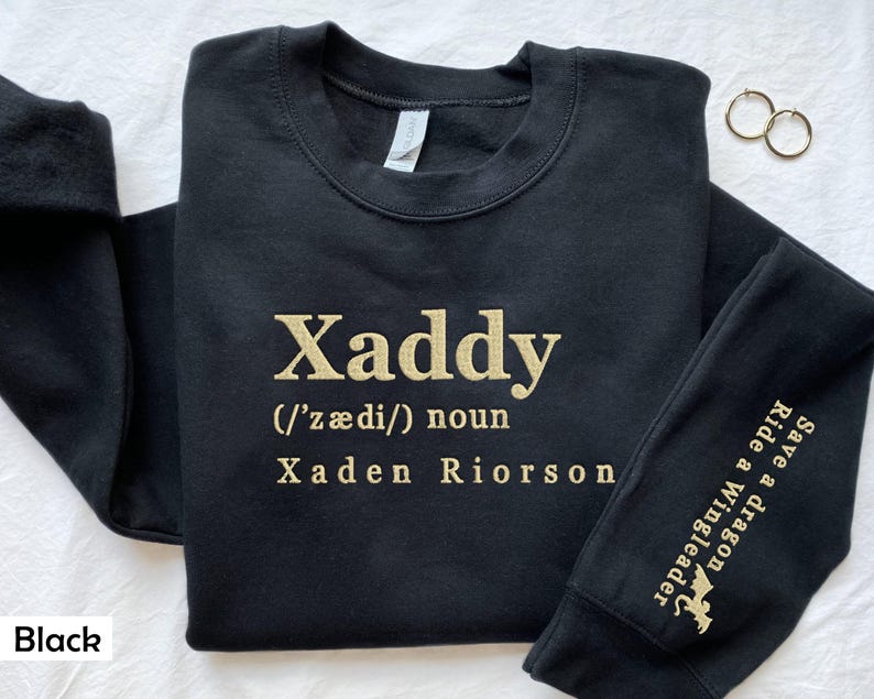 May include: Black sweatshirt with gold lettering that reads "Xaddy (/ˈz&aelig;di/) noun Xaden Riorson". The right sleeve has a gold dragon graphic and the text "Ride a Wingleader Save a dragon".