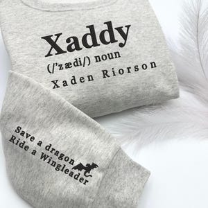 May include: A gray sweatshirt with the text "Xaddy (/ˈz&aelig;di/) noun Xaden Riorson" on the front and "Save a dragon Ride a Wingleader" on the sleeve. The sweatshirt is labeled "Sport Grey".