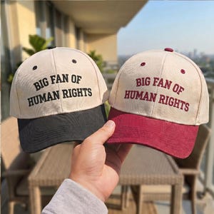 Pode incluir: Dois bonés de beisebol bege com o texto "BIG FAN OF HUMAN RIGHTS". Um boné tem a aba preta, o outro bordô. O texto é preto e bordô.