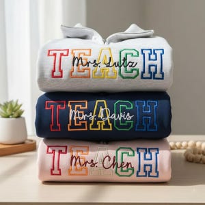 May include: Three folded sweatshirts, each embroidered with "TEACH" in colorful block letters and a teacher's name. The top sweatshirt is light gray, the middle is navy blue, and the bottom is pink. A succulent plant in a white pot is visible.