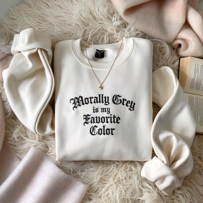 May include: Cream-colored sweatshirt with the phrase "Morally Grey is my Favorite Color" in black script. A gold necklace with a small pendant rests on the shirt. The sleeves are folded, and the sweatshirt is on a fluffy, white surface.
