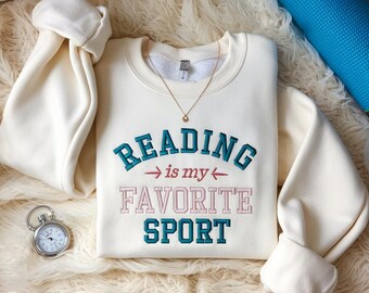 Könnte beinhalten: Cremefarbenes Sweatshirt mit dem gestickten Text "READING is my FAVORITE SPORT" in Türkis und Rosa. Eine goldene Halskette und eine Stoppuhr sind ebenfalls sichtbar. Die Ärmel des Sweatshirts sind gefaltet, und eine blaue Yogamatte ist im Hintergrund.