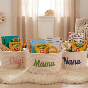 May include: Three cream-colored rope baskets labeled "Gigi," "Mama," and "Nana." Each basket contains school supplies, including Crayola crayons, notebooks, and books. The baskets sit on a fluffy white rug.