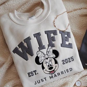Puede incluir: Sudadera color crema con la palabra "WIFE" en gris, con un gr&aacute;fico de Minnie Mouse y el texto "EST. JUST MARRIED 2025". Tambi&eacute;n se ven un collar dorado y relojes.