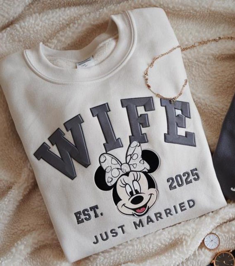 Puede incluir: Sudadera color crema con la palabra "WIFE" en gris, con un gr&aacute;fico de Minnie Mouse y el texto "EST. JUST MARRIED 2025". Tambi&eacute;n se ven un collar dorado y relojes.