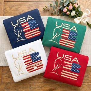 Puede incluir: Cuatro sudaderas dobladas en azul marino, verde, blanco y rojo. Cada una presenta el texto bordado USA 26, un gráfico de trofeo estilizado y un diseño rectangular que incorpora las estrellas y rayas de la bandera estadounidense en rojo, blanco y azul.