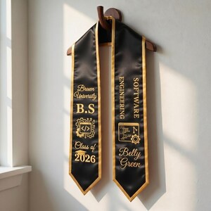 May include: Black graduation sash with gold trim and lettering. The sash reads "Brown University B.S" and "Software Engineering" with the name "Belly Green" and "Class of 2026". The sash is hanging on a wooden hanger.