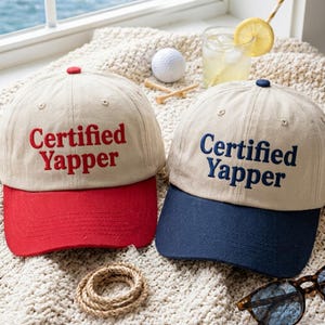 May include: Two baseball caps with beige crowns and embroidered text reading Certified Yapper. One cap features a red brim and red lettering, while the other has a navy blue brim and navy blue lettering. Both caps have a button on top.