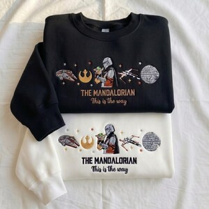 May include: A black sweatshirt and a white sweatshirt, each featuring embroidered Star Wars designs including a Mandalorian character, a small green creature, a spaceship, a rebel symbol, and a Death Star, with the text THE MANDALORIAN This is the way.