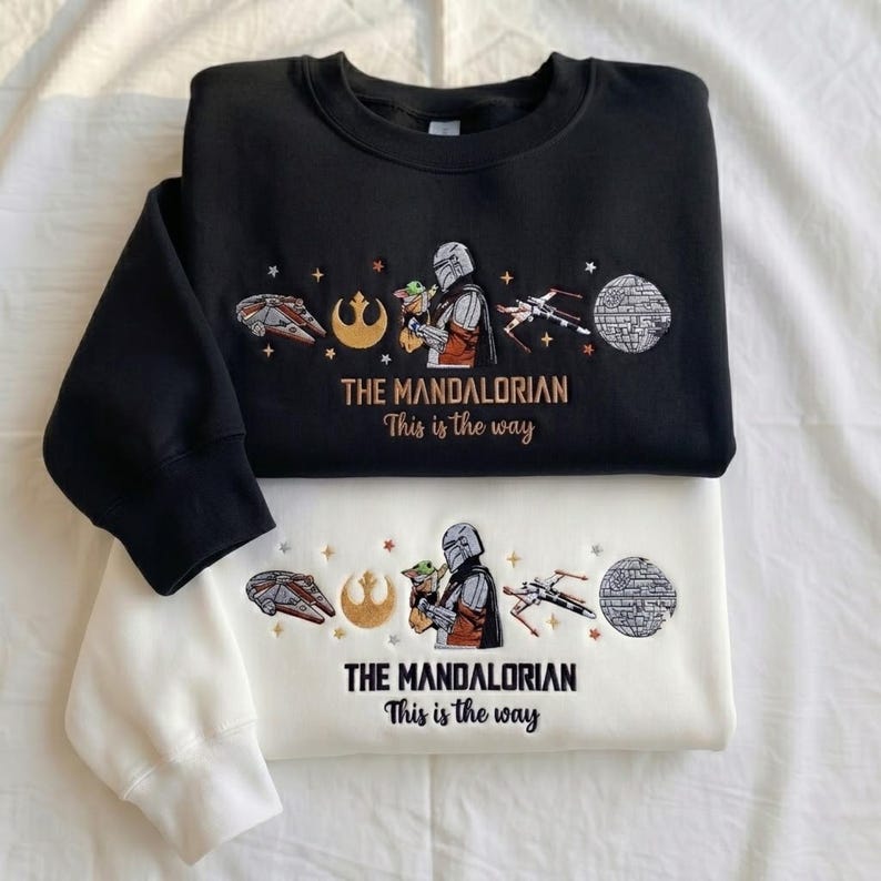 May include: A black sweatshirt and a white sweatshirt, each featuring embroidered Star Wars designs including a Mandalorian character, a small green creature, a spaceship, a rebel symbol, and a Death Star, with the text THE MANDALORIAN This is the way.