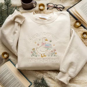 Può includere: Felpa color crema con design ricamato. Il design presenta un'oca bianca che trasporta libri, con la scritta "OOPS, I BOUGHT ANOTHER BOOK". Il design include fiori e stelle. La felpa ha girocollo e maniche lunghe.