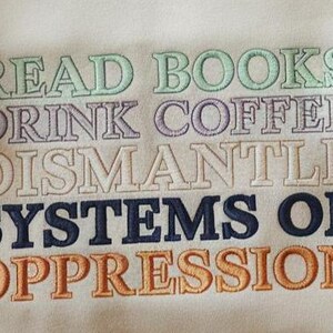 May include: Cream-colored sweatshirt with embroidered text in various colors. The text reads "READ BOOKS DRINK COFFEE DISMANTLE SYSTEMS OF OPPRESSION". The embroidery is in shades of green, purple, white, navy blue, and orange.
