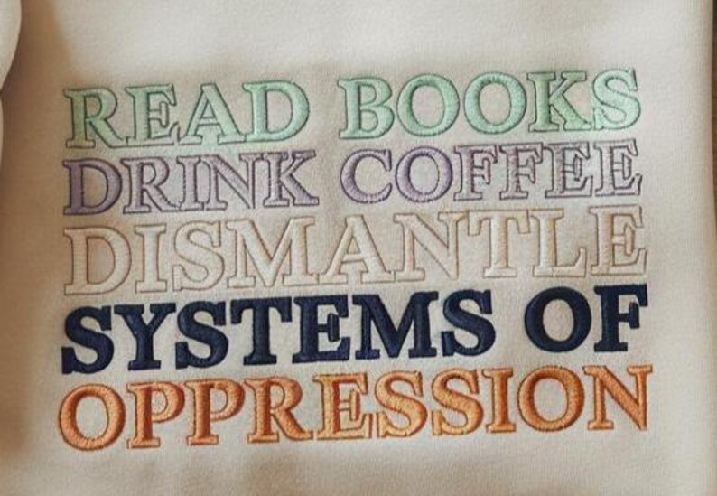May include: Cream-colored sweatshirt with embroidered text in various colors. The text reads "READ BOOKS DRINK COFFEE DISMANTLE SYSTEMS OF OPPRESSION". The embroidery is in shades of green, purple, white, navy blue, and orange.