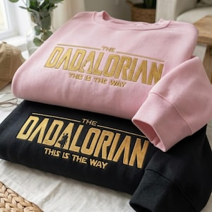 Może przedstawiać: Dwie bluzy, jedna r&oacute;żowa i jedna czarna, są ułożone jedna na drugiej. R&oacute;żowa bluza ma złoty napis "THE DADALORIAN THIS IS THE WAY". Czarna bluza ma ten sam złoty napis. Widoczne są rękawy.