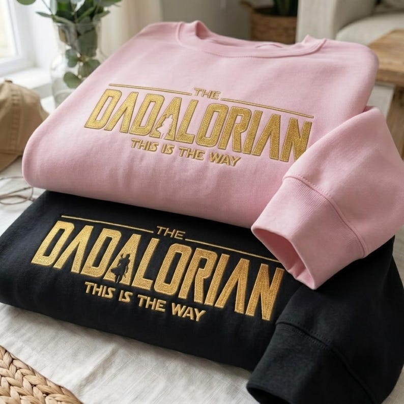Może przedstawiać: Dwie bluzy, jedna r&oacute;żowa i jedna czarna, są ułożone jedna na drugiej. R&oacute;żowa bluza ma złoty napis "THE DADALORIAN THIS IS THE WAY". Czarna bluza ma ten sam złoty napis. Widoczne są rękawy.