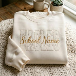 Könnte beinhalten: Cremefarbenes Sweatshirt mit dem Wort "EAGLES" in Blockbuchstaben und "School Name" in Schreibschrift gestickt. Das Sweatshirt liegt gefaltet auf einer strukturierten Oberfläche, im Hintergrund sind eine Tasse und eine kleine Pflanze zu sehen. Das Design ist goldfarben.