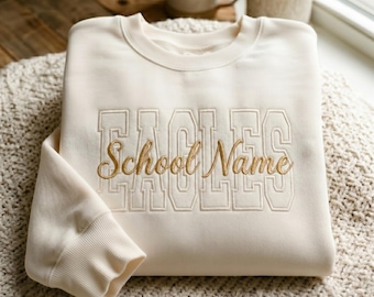 May include: Cream-colored sweatshirt with the word "EAGLES" embroidered in block letters and "School Name" in cursive script. The sweatshirt is folded on a textured surface, with a mug and small plant visible in the background. The design is in a gold color.
