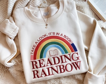 May include: A cream-colored crewneck sweatshirt featuring an embroidered design of a rainbow with a small book icon in the center. The text reads "TAKE A LOOK, IT'S IN A BOOK!" in an arc above the rainbow and "READING RAINBOW" in large red letters.