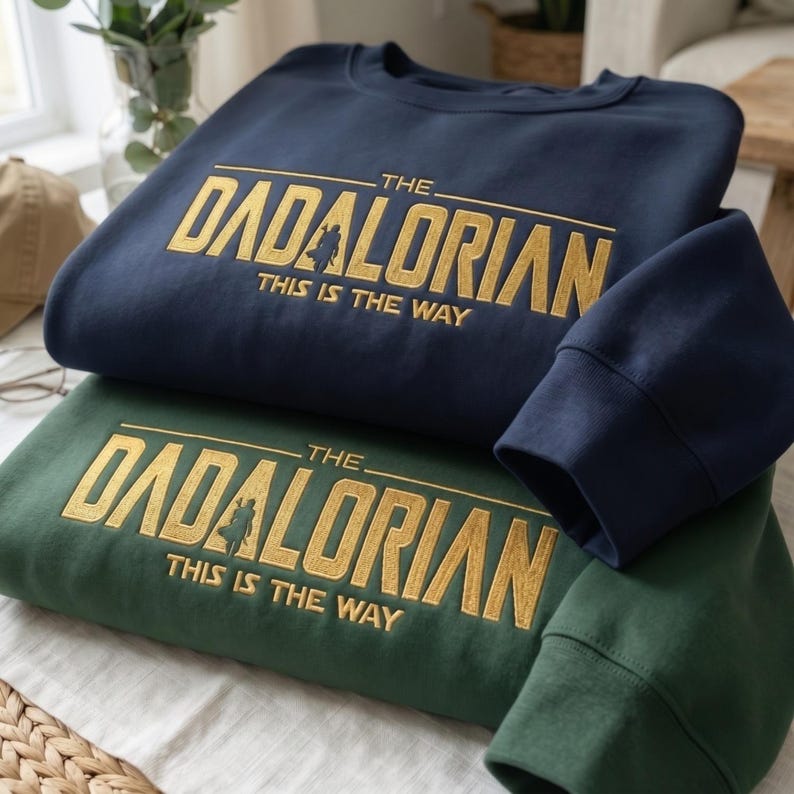 Może przedstawiać: Dwie bluzy, jedna granatowa i jedna zielona, są ułożone jedna na drugiej. Granatowa bluza ma złoty napis "THE DADALORIAN THIS IS THE WAY". Zielona bluza ma ten sam napis. Widoczne są rękawy granatowej bluzy.