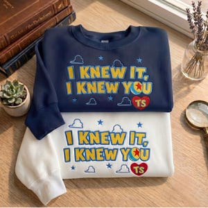 May include: A navy blue sweatshirt and a white sweatshirt, each featuring embroidered text that reads I KNEW IT, I KNEW YOU with a ball graphic, a red heart with the letters TS, and small blue cloud and star shapes scattered across the front.