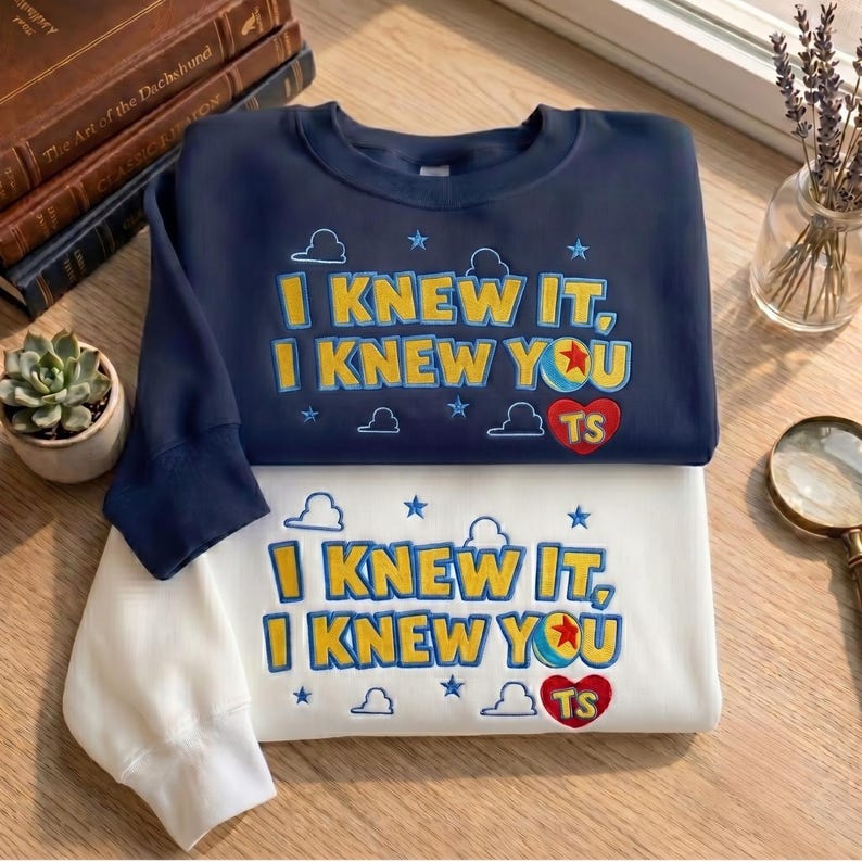 May include: A navy blue sweatshirt and a white sweatshirt, each featuring embroidered text that reads I KNEW IT, I KNEW YOU with a ball graphic, a red heart with the letters TS, and small blue cloud and star shapes scattered across the front.