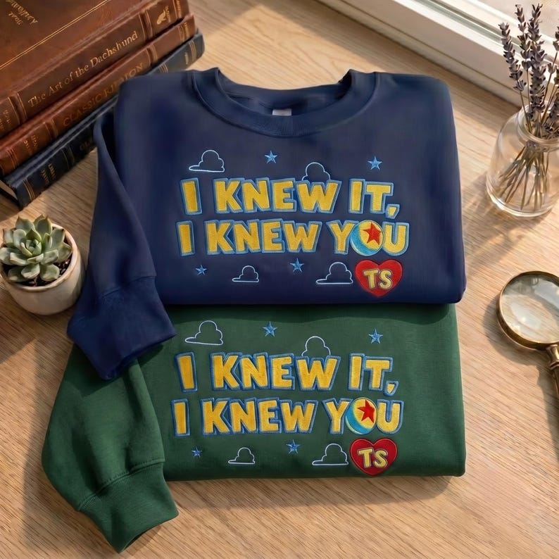 May include: A navy blue sweatshirt and a dark green sweatshirt, each featuring the yellow text "I KNEW IT, I KNEW YOU" with a colorful ball icon, a red heart with "TS" inside, and small blue cloud and star outlines embroidered on the front.