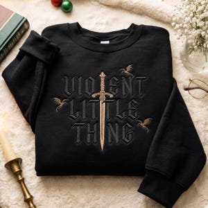 Könnte beinhalten: Ein schwarzes Sweatshirt mit Rundhalsausschnitt und goldener Stickerei, die ein zentrales Schwert zeigt, welches den Text "VIOLENT LITTLE THING" in gotischer Schrift teilt. Drei kleine goldene Drachen sind um das Motiv angeordnet.