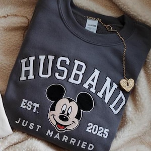 Puede incluir: Una sudadera gris carb&oacute;n con la palabra "HUSBAND" en letras blancas, junto con "EST. JUST MARRIED 2025" y un dibujo de Mickey Mouse. Un colgante dorado en forma de coraz&oacute;n cuelga del cuello.