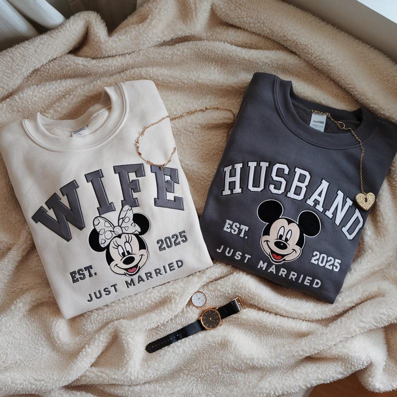 Puede incluir: Dos sudaderas a juego, una crema y otra gris carb&oacute;n, con las palabras "WIFE" y "HUSBAND" en negrita. Cada una presenta un dise&ntilde;o de Mickey Mouse y el texto "EST. JUST MARRIED 2025". Tambi&eacute;n se ven collares dorados y un reloj.