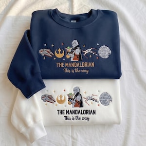 May include: A navy blue and a white crewneck sweatshirt, each featuring embroidered designs of a Mandalorian character holding a small green creature, a spaceship, a starfighter, a circular space station, and the text THE MANDALORIAN This is the way.