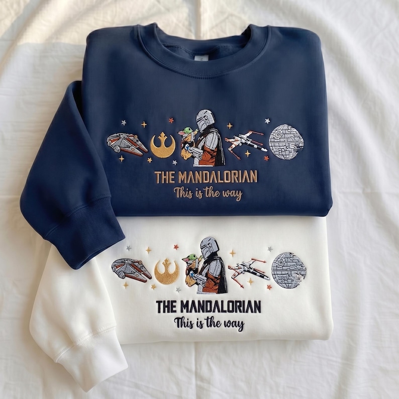 May include: A navy blue and a white crewneck sweatshirt, each featuring embroidered designs of a Mandalorian character holding a small green creature, a spaceship, a starfighter, a circular space station, and the text THE MANDALORIAN This is the way.
