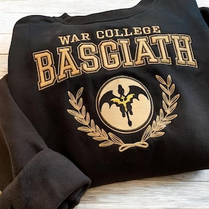 May include: Black sweatshirt with gold embroidery. The embroidery says "War College Basciath" and features a winged dragon inside a circle with a laurel wreath.
