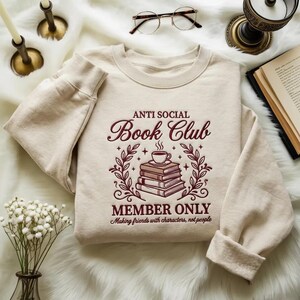 May include: A cream-coloured sweatshirt featuring the burgundy text "ANTI SOCIAL Book Club". The design includes a stack of books topped with a coffee cup, framed by leaves. The phrases "MEMBER ONLY" and "Making friends with characters, not people" are also present.