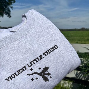 Puede incluir: Una sudadera gris con el texto "VIOLENT LITTLE THING" impreso en la parte delantera. La sudadera tambi&eacute;n presenta un peque&ntilde;o gr&aacute;fico negro de un drag&oacute;n con estrellas.