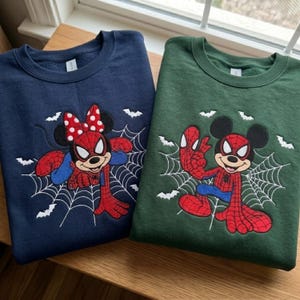 May include: A navy blue sweatshirt and a dark green sweatshirt, each featuring an embroidered graphic of a cartoon mouse character dressed in a red and blue spider-themed costume, positioned on a white spider web with small white bats in the background.