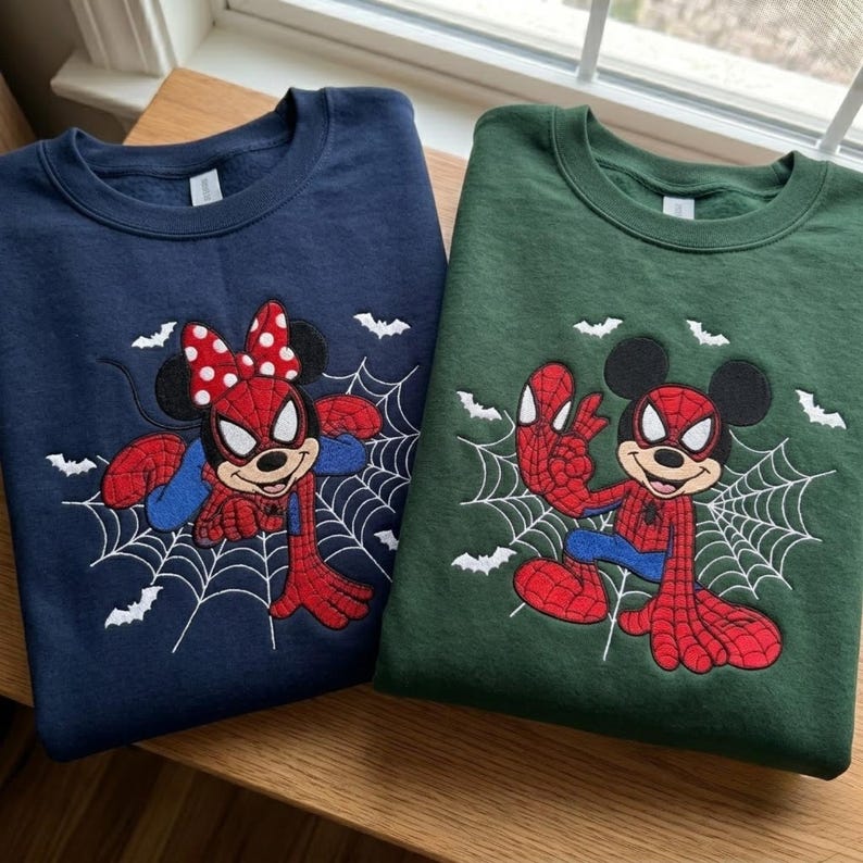 May include: A navy blue sweatshirt and a dark green sweatshirt, each featuring an embroidered graphic of a cartoon mouse character dressed in a red and blue spider-themed costume, positioned on a white spider web with small white bats in the background.