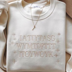 May include: Cream-colored sweatshirt with embroidered text and starburst designs. A gold-colored necklace with a pendant rests on the folded garment. The text on the sweatshirt is in a light brown color.
