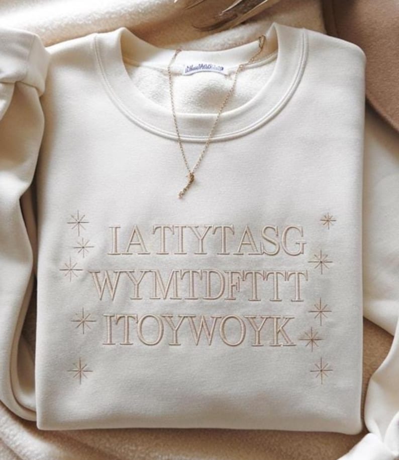 May include: Cream-colored sweatshirt with embroidered text and starburst designs. A gold-colored necklace with a pendant rests on the folded garment. The text on the sweatshirt is in a light brown color.