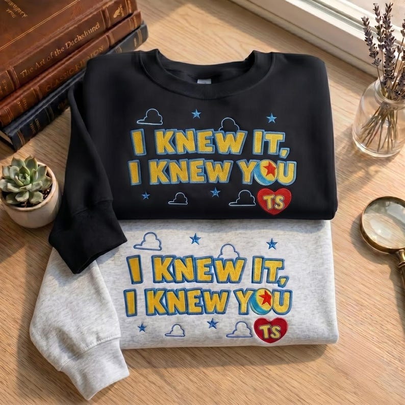 May include: A black sweatshirt and a light gray sweatshirt, each featuring embroidered text that reads I KNEW IT, I KNEW YOU with a colorful ball icon and a red heart containing the letters TS, surrounded by small blue stars and cloud outlines.