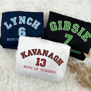 Puede incluir: Tres sudaderas blancas con texto y números de diferentes colores. La primera sudadera tiene texto azul que dice "Lynch 6 Boys of", la segunda sudadera tiene texto verde que dice "Gibsie 7 Mmen", y la tercera sudadera tiene texto rojo que dice "Kavanagh 13 Boys of Tommen".
