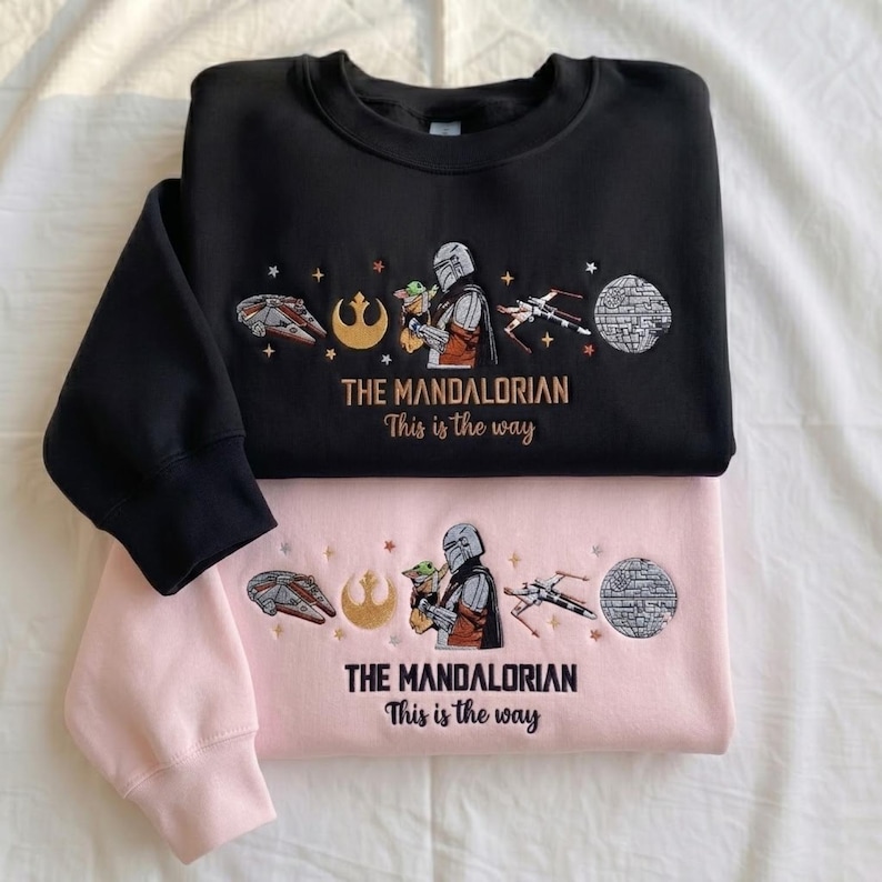 May include: A black sweatshirt and a light pink sweatshirt, each featuring embroidered designs of a Mandalorian character holding a small creature, a spaceship, a starfighter, a Death Star, and the text THE MANDALORIAN This is the way.