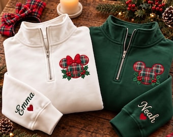 May include: A white and a dark green quarter-zip sweatshirt, each featuring an embroidered plaid mouse-ear icon with holly berries. The white sweatshirt sleeve is embroidered with the name Emma and a red heart, while the green sleeve features the name Noah and a red heart.