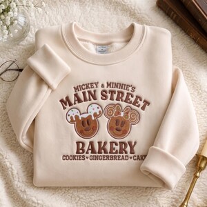 May include: A cream-colored crewneck sweatshirt featuring brown embroidered text that reads Mickey & Minnie's Main Street Bakery, Cookies, Gingerbread, Cake. The design includes two gingerbread-style character faces with icing details.