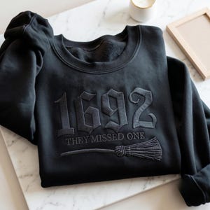 Könnte beinhalten: Ein schwarzes Sweatshirt mit Rundhalsausschnitt und dunkelgrauer Stickerei auf der Brust. Das Design zeigt die großen Zahlen 1692, darunter den Text "THEY MISSED ONE" sowie eine Grafik eines Besenstiels, die unter dem Text in passendem Garn gestickt ist.